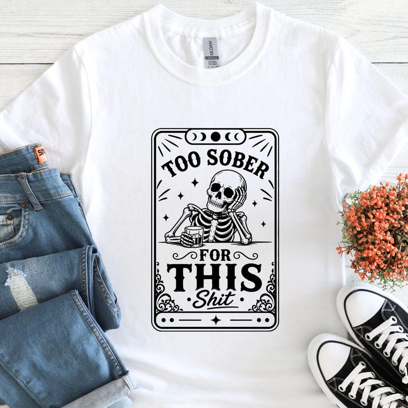 K&ouml;nnte beinhalten: Wei&szlig;es T-Shirt mit einer schwarz-wei&szlig;en Grafik eines Skeletts, das ein Getr&auml;nk h&auml;lt, mit dem Text "TOO SOBER FOR THIS Shit". Das Shirt wird mit blauen Jeans und schwarz-wei&szlig;en Turnschuhen gezeigt.