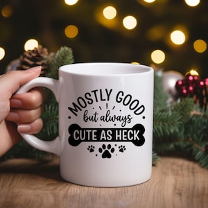 May include: White ceramic mug with the text "Mostly Good but always Cute as Heck" in black. The design includes a bone and paw prints. The mug is held by a hand, with a blurred background of Christmas decorations.