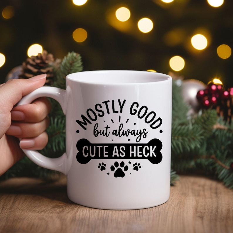 May include: White ceramic mug with the text "Mostly Good but always Cute as Heck" in black. The design includes a bone and paw prints. The mug is held by a hand, with a blurred background of Christmas decorations.