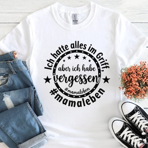 May include: White t-shirt with a black circular design that reads "Ich hatte alles im Griff, aber ich habe vergessen #mamaleben". The shirt is paired with blue jeans and black and white sneakers.