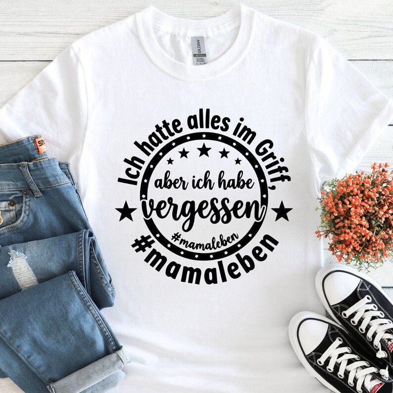 May include: White t-shirt with a black circular design that reads "Ich hatte alles im Griff, aber ich habe vergessen #mamaleben". The shirt is paired with blue jeans and black and white sneakers.