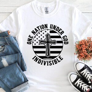 May include: White t-shirt with a black graphic featuring a cross, stars, and stripes design. The text reads "ONE NATION UNDER GOD INDIVISIBLE". The shirt is paired with blue jeans and black and white sneakers.