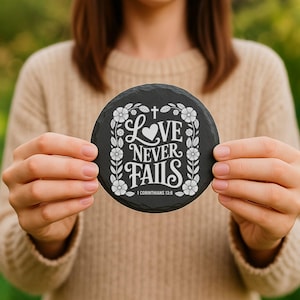 Puede incluir: Un posavasos redondo de pizarra negra con detalles florales blancos y el texto "Love Never Fails" en una fuente decorativa. Tambi&eacute;n incluye el vers&iacute;culo b&iacute;blico "1 Corintios 13:8".