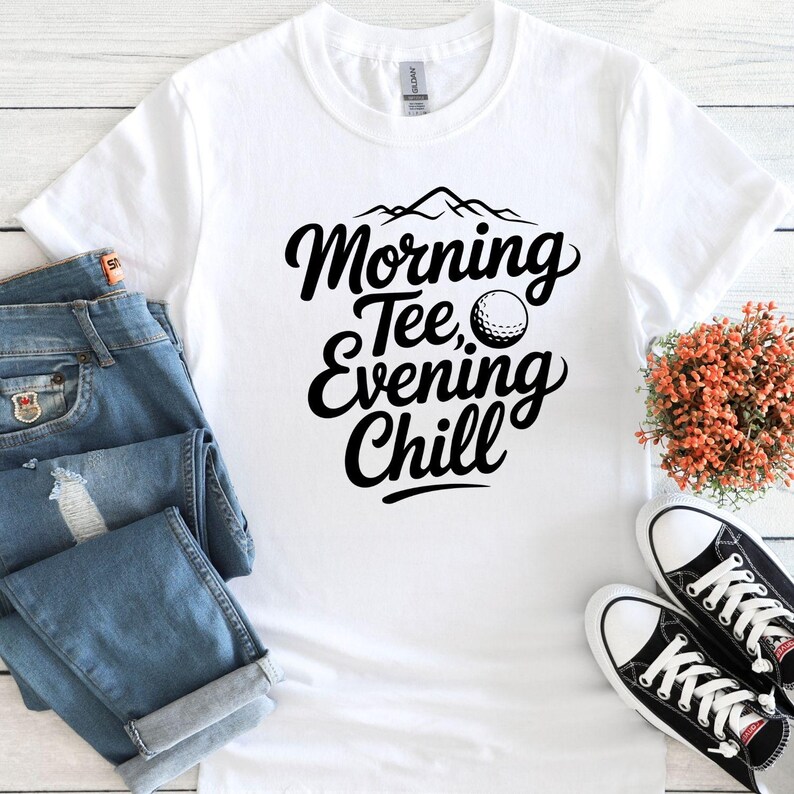 May include: White t-shirt with the text "Morning Tee, Evening Chill" in black script, featuring a golf ball graphic. The shirt is paired with blue jeans and black and white sneakers. A small bouquet of orange flowers is also present.