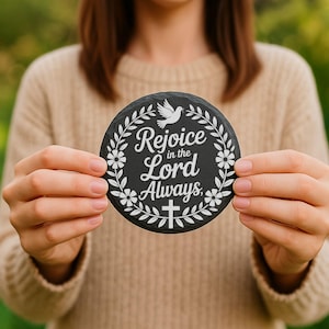 Puede incluir: Un posavasos redondo y negro con texto blanco que dice "Rejoice in the Lord Always". El posavasos presenta una paloma blanca, detalles florales y una cruz. El posavasos est&aacute; sostenido por una persona.