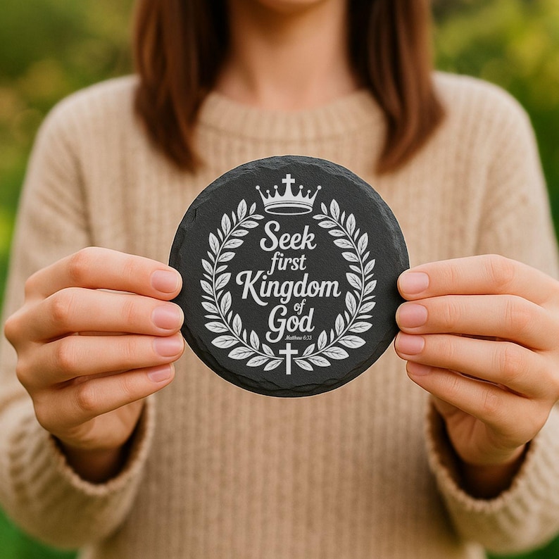 Puede incluir: Un posavasos redondo de pizarra negra con el texto blanco "Seek first Kingdom of God", rodeado de una corona de laurel, una corona y una cruz. El posavasos se sostiene frente a un fondo borroso.
