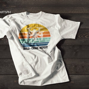 Peut inclure: T-shirt blanc avec un graphique de style vintage d'un drone volant devant un coucher de soleil. Le texte "BUILD - CRASH - REPEAT" est imprimé sous le graphique.