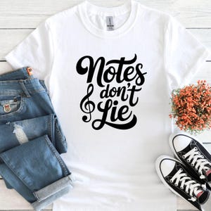 May include: White t-shirt with the text "Notes don't Lie" in black script, featuring a treble clef. The shirt is paired with blue jeans and black and white sneakers, suggesting a music-themed style.