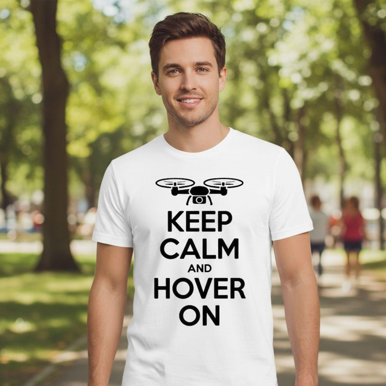 May include: White t-shirt with a black drone graphic and the words "KEEP CALM AND HOVER ON". The shirt is made of a soft material and is suitable for casual wear. The design features a drone illustration above the text.
