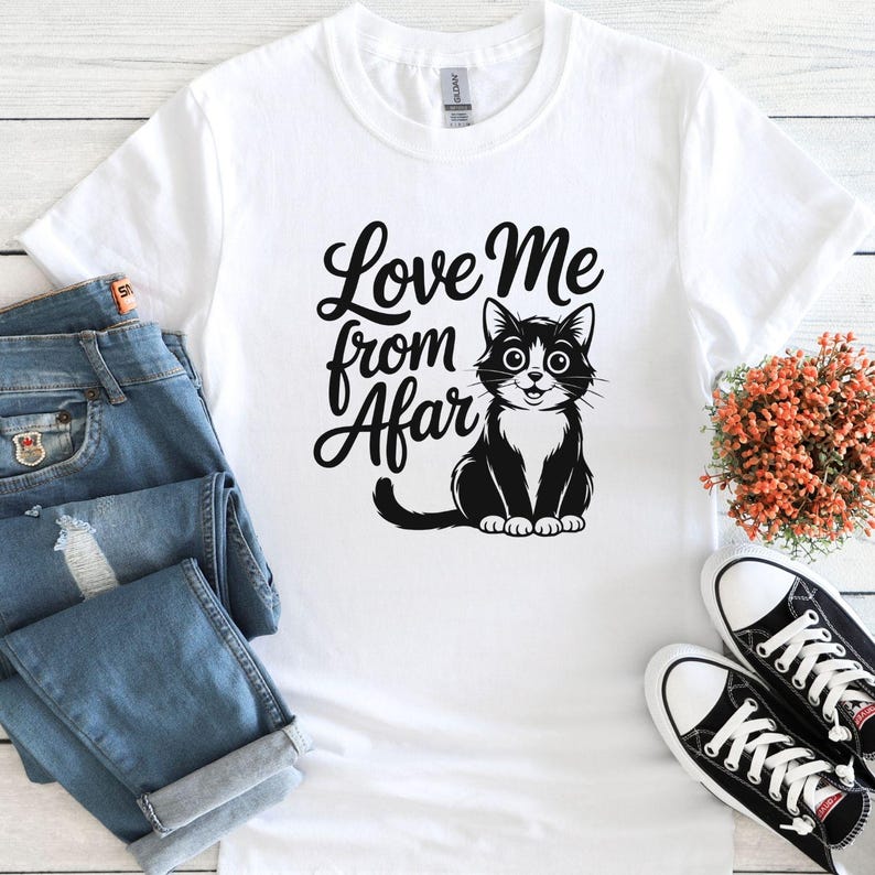 May include: White t-shirt with the text "Love Me from Afar" and a black and white cat illustration. The cat has large eyes and a smiling expression. The shirt is paired with blue jeans and black and white sneakers.