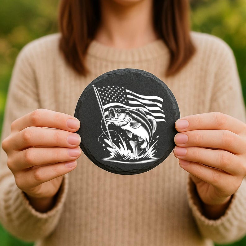 May include: A round black slate coaster featuring a white graphic of a leaping fish and the American flag. The design includes stars, stripes, and a fish jumping out of the water. The coaster is held up by a person.