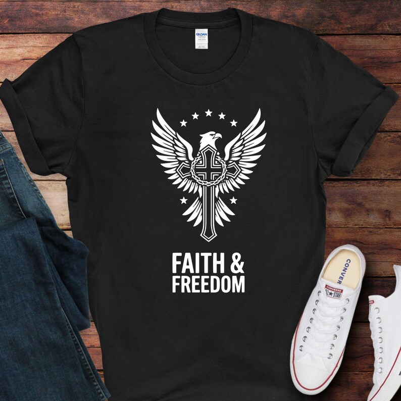 May include: Black t-shirt featuring a white graphic of an eagle with outstretched wings, a cross, and the text "FAITH & FREEDOM". The design includes stars and a decorative border. The shirt is displayed on a wooden surface.