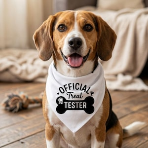 May include: A happy beagle dog wearing a white bandana with the text "OFFICIAL TREAT TESTER" in black. The dog has brown and white fur and is sitting on a wooden floor.