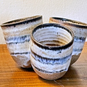 May include: Three handmade ceramic cups with a unique, layered design. The cups feature a blend of brown, white, and blue hues, with a dark brown rim. The cups are displayed on a wooden surface.