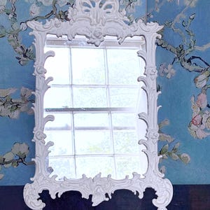 May include: A white, ornate mirror with a decorative frame. The mirror has a rectangular shape with a detailed, scrollwork design. The background features a blue wall with a floral painting.
