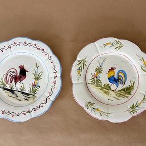 May include: Two decorative ceramic plates with scalloped edges. Each plate features a hand-painted rooster design in vibrant colors. One plate has a blue border, the other a pink border.
