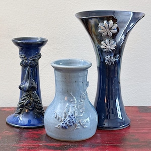 May include: Three ceramic vases in shades of blue. The tallest vase is dark blue with embossed flowers. The middle vase is light blue with grape and leaf detailing. The third vase is a dark blue candlestick holder with a decorative design.