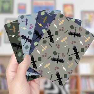 Op de afbeelding: Een hand houdt een waaier van speelkaarten met een herhalend patroon van zwarte draken, bliksemschichten, zwaarden en andere fantasie-elementen. De kaarten hebben een verscheidenheid aan achtergrondkleuren, waaronder groen, blauw en grijs.