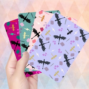 May include: A hand holds four rectangular cards with dragon and fantasy-themed patterns. The cards feature black dragons, swords, lightning bolts, and other fantasy elements on backgrounds of pink, teal, purple, and magenta.