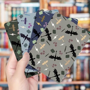 Op de afbeelding: Een hand die vier kaarten met patronen vasthoudt. Elke kaart heeft een herhalend patroon van zwarte draken, bliksemschichten en andere fantasie-elementen in tinten groen, blauw en grijs. De achtergrond is een vage boekenkast.