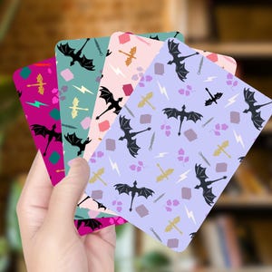 May include: A hand holds a stack of colorful cards with a fantasy theme. Each card features a repeating pattern of black dragons, swords, lightning bolts, and other fantasy elements on backgrounds of pink, purple, teal, and magenta.