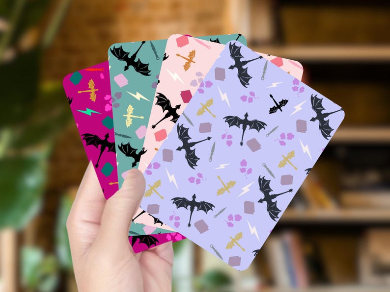 May include: A hand holds a stack of colorful cards with a fantasy theme. Each card features a repeating pattern of black dragons, swords, lightning bolts, and other fantasy elements on backgrounds of pink, purple, teal, and magenta.