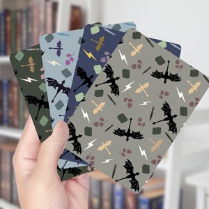 Op de afbeelding: Een hand houdt een waaier van vijf rechthoekige kaarten met draakthema's vast. Elke kaart heeft een ander kleurenschema, waaronder groen, blauw en grijs, met herhaalde illustraties van draken, bliksemschichten en zwaarden. De kaarten zijn waarschijnlijk bladwijzers of decoratieve items.