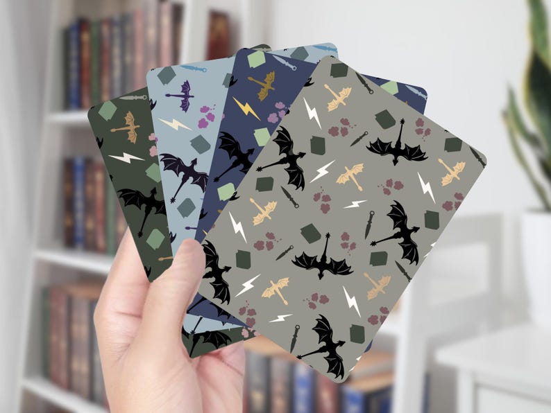 Op de afbeelding: Een hand houdt een waaier van vijf rechthoekige kaarten met draakthema's vast. Elke kaart heeft een ander kleurenschema, waaronder groen, blauw en grijs, met herhaalde illustraties van draken, bliksemschichten en zwaarden. De kaarten zijn waarschijnlijk bladwijzers of decoratieve items.