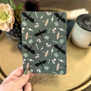 Op de afbeelding: Een rechthoekig notitieboek met een donkergroene achtergrond en een herhalend patroon van zwarte draken, bliksemschichten, zwaarden en roze bloemaccenten. Het notitieboek ligt op een gouden dienblad.