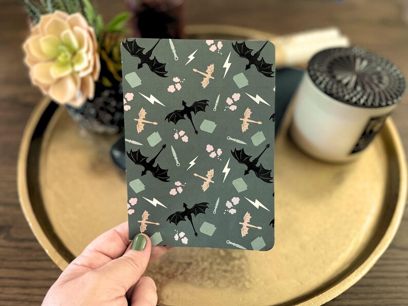 Op de afbeelding: Een rechthoekig notitieboek met een donkergroene achtergrond en een herhalend patroon van zwarte draken, bliksemschichten, zwaarden en roze bloemaccenten. Het notitieboek ligt op een gouden dienblad.