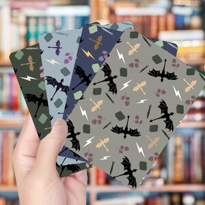 May include: A hand holding a fan of four patterned cards. Each card features a repeating pattern of black dragons, lightning bolts, and other fantasy-themed elements in shades of green, blue, and gray. The background is a blurred bookshelf.