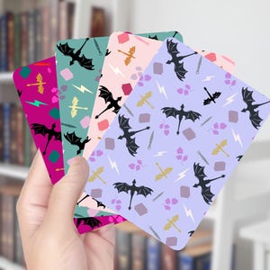 May include: A hand holds a stack of colorful cards with dragon and fantasy-themed patterns. Each card features a different background color: magenta, teal, pink, and lavender. The patterns include black dragons, swords, lightning bolts, and other fantasy elements.