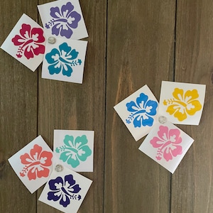 May include: Assortment of eight hibiscus flower stickers in various colors, including pink, purple, blue, teal, and yellow. The stickers are on a wood background.