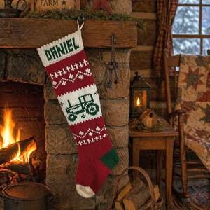 May include: A red and white knit Christmas stocking featuring a green tractor graphic and the name DANIEL knitted in green at the top. The stocking has a ribbed cuff, a green heel and toe section, and a white toe tip, with a hanging loop at the top.