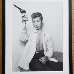 May include: Black and white framed print featuring a man holding a gun. The man is wearing a light-colored shirt and dark pants. The image is a portrait, with the man looking towards the viewer.