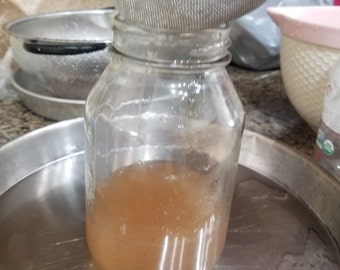 May include: A clear glass mason jar containing a light brown liquid, positioned beneath a fine-mesh metal strainer. The jar has a wide mouth and a threaded rim, and the liquid inside appears translucent with a smooth, consistent texture throughout.