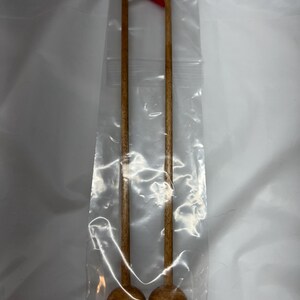 May include: Two long, slender wooden sticks with rounded, dome-shaped bases at the bottom. The sticks are light brown in color and are enclosed within a clear plastic bag. At the top of the sticks, small white and red triangular fabric pieces are visible.
