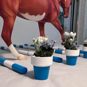 May include: A brown and white horse figurine with a raised leg, positioned above blue and white striped poles and small blue flower pots with white rims. The pots contain small white and purple flowers.