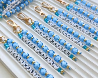 May include: A collection of beaded keychains featuring light blue faceted beads, small gold-tone spacer beads, and white letter beads spelling TICKETMASTER. Each keychain is finished with a gold-tone lobster clasp for attachment to bags or keys.