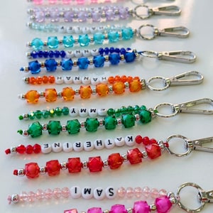 May include: A collection of colorful beaded keychains featuring faceted plastic beads in shades of pink, purple, blue, orange, green, and red. Each keychain includes white letter beads spelling names like Emmy, Hannah, Katherine, and Sami.