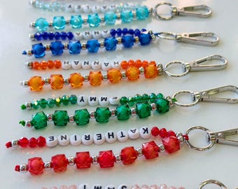 May include: A collection of colorful beaded keychains featuring faceted plastic beads in shades of pink, purple, blue, orange, green, and red. Each keychain includes white letter beads spelling names like Emmy, Hannah, Katherine, and Sami.