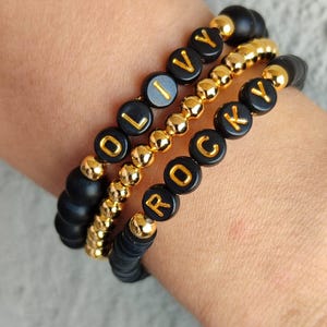 May include: Three stacked bracelets featuring matte black beads and polished gold-tone metallic beads. One bracelet displays the name OLIVY in gold letters on black beads, while another features the name ROCKY in gold letters on black beads.