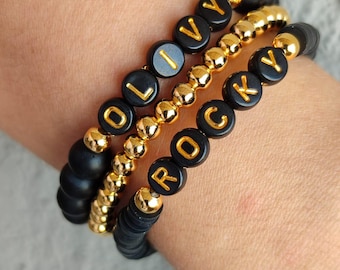 May include: Three stacked bracelets featuring matte black beads and polished gold-tone metallic beads. One bracelet displays the name OLIVY in gold letters on black beads, while another features the name ROCKY in gold letters on black beads.