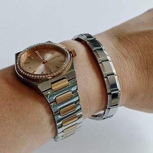 May include: A silver and gold-tone watch with a round, rose-gold colored face and a bezel encrusted with small crystals. A silver bracelet with rectangular links is also shown.