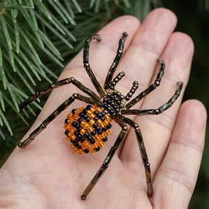 May include: A beaded spider with a black and orange abdomen, crafted with black and gold beads. The spider is sitting on a person's hand.