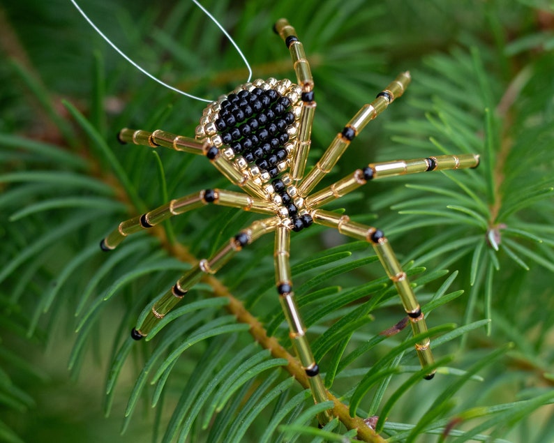 May include: A gold and black beaded spider ornament hanging from a white string. The spider has a black body and gold legs with black accents.