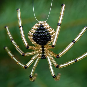 May include: A beaded spider ornament with a black and gold color scheme. The spider has eight legs and a round body with a black bead for an eye.