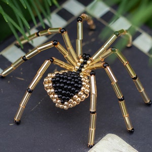 May include: A beaded spider with a black and gold body and eight long legs. The spider is made of small beads and is a decorative piece.