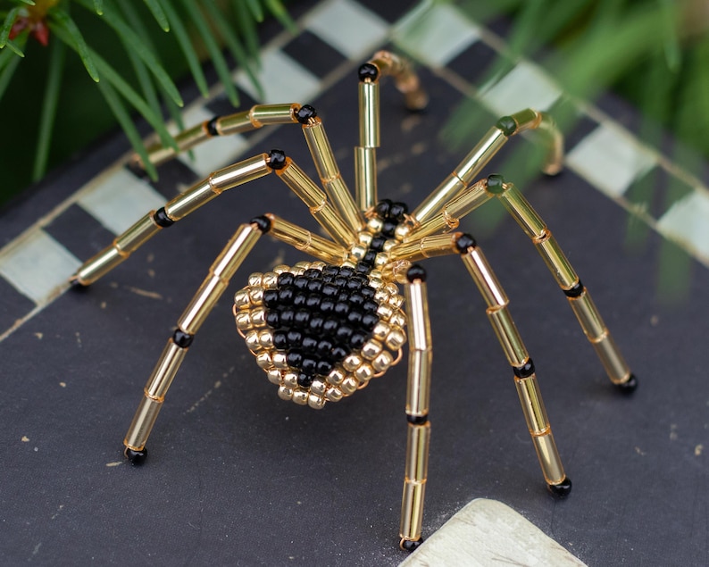 May include: A beaded spider with a black and gold body and eight long legs. The spider is made of small beads and is a decorative piece.