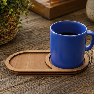 Pode incluir: Uma caneca de cer&acirc;mica azul s&oacute;lida com al&ccedil;a est&aacute; sobre uma bandeja de madeira marrom clara. A bandeja tem um formato oval com um recorte circular projetado para segurar a base da caneca com seguran&ccedil;a, deixando uma superf&iacute;cie plana ao lado.